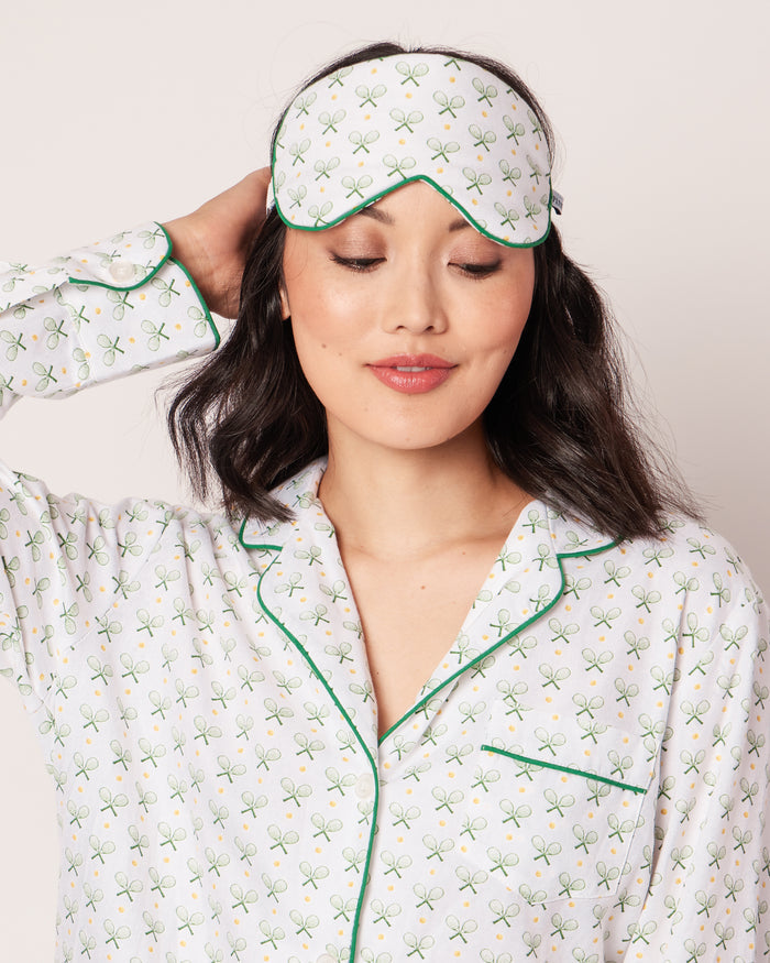 A person wearing tennis print pajamas adjusts a Petite Plume Adults Sleep Mask in Match Point on their forehead. The white mask with green detailing complements the plain light-colored background, and the individuals long dark hair completes this cozy yet stylish ensemble.