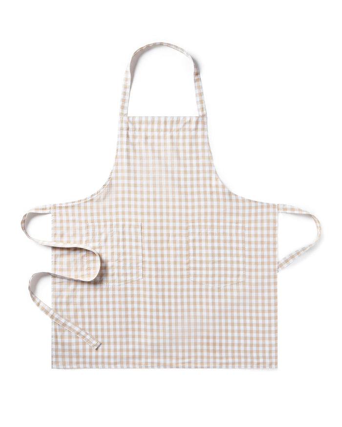 The Adult's Twill Apron in Camel Gingham features a beige and white checkered pattern, a neck loop, front pockets, and waist ties, shown laid flat on a white background.