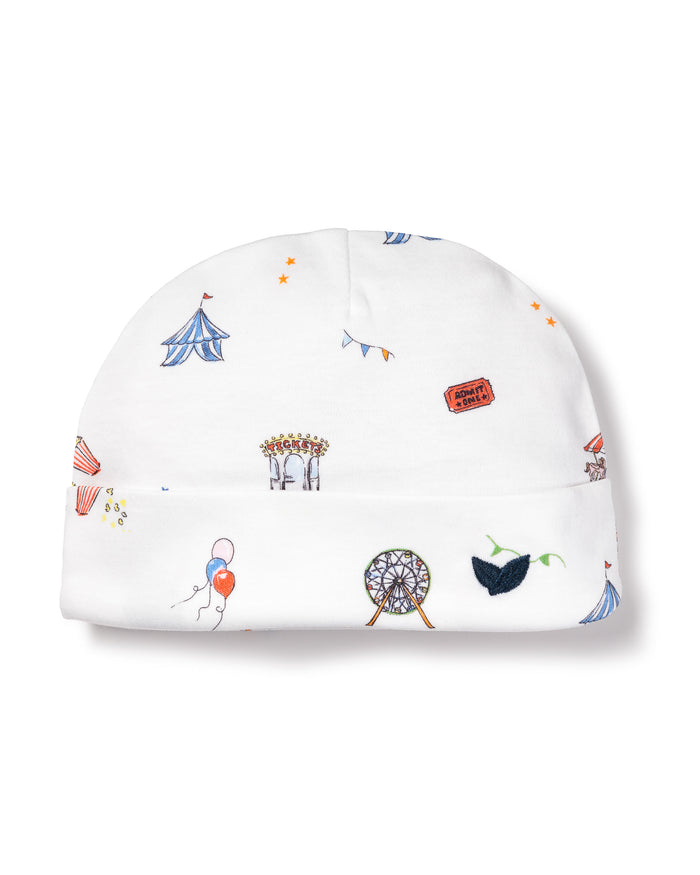 The Babys Pima Hat in Carnival Memories by Petite Plume is made from soft Peruvian Pima cotton, featuring playful circus patterns like tents, carousels, Ferris wheels, balloons, green leaves, and orange tickets scattered across the fabric.