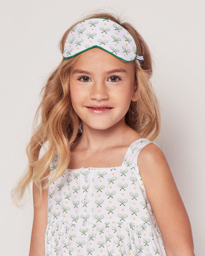 A young tennis enthusiast beams in a white sleeveless dress with racket prints, her light brown hair framing her face. She wears the Kids Sleep Mask in Match Point by Petite Plume on her head, and the plain background emphasizes her cheerful expression.