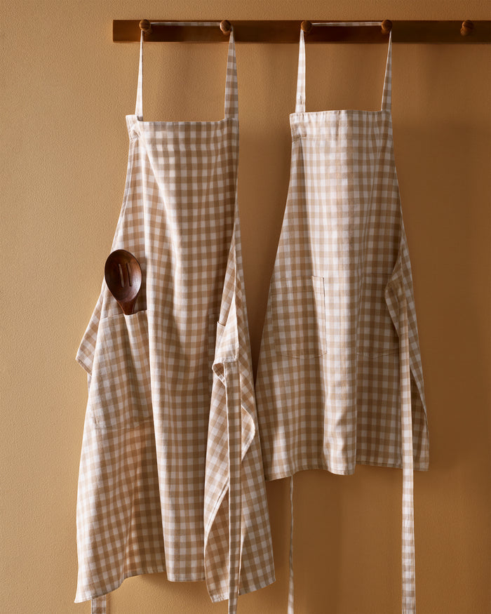 The Kid's Twill Apron in Camel Gingham, featuring a beige and white gingham pattern, two front pockets, and long waist ties, is displayed on a white background.