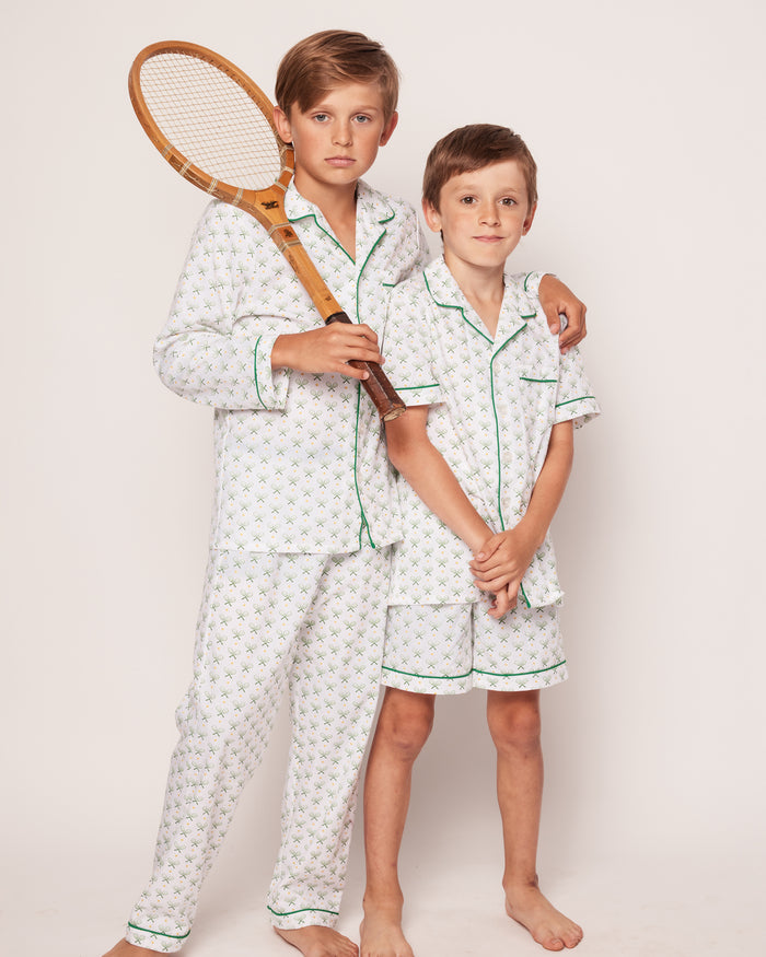 Two young boys model Petite Plumes Kids Twill Pajama Set in Match Point. The older boy, holding a wooden tennis racket that suggests their passion for the sport, affectionately wraps an arm around the younger boy as they stand barefoot against a simple backdrop.