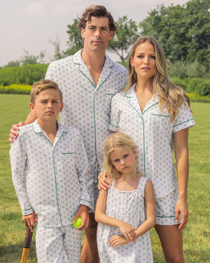 A family of four stands on a grassy field wearing matching Petite Plume sleepwear featuring a subtle pattern. The boy holds a tennis ball, hinting at his Kids Twill Pajama Set in Match Point. Trees and a cloudy sky provide the serene backdrop.