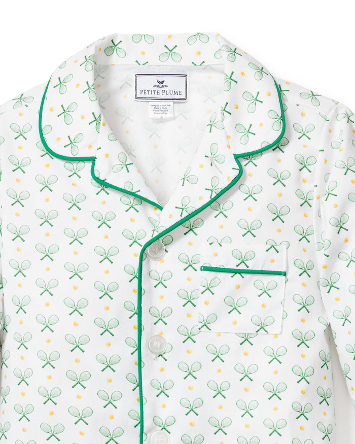 The Kids Twill Pajama Set in Match Point by Petite Plume features a white top with a green tennis racket pattern, yellow accents, and green piping on the collar, button line, and pocket edge. A Petite Plume label adorns the collar for a luxurious sleepwear charm.