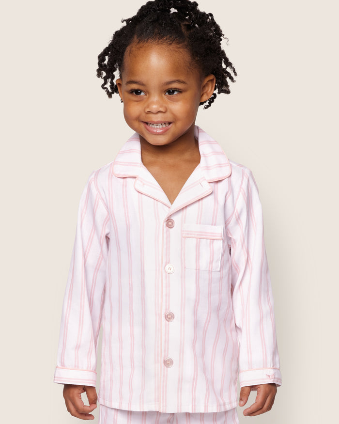 A young child with curly hair smiles while wearing Petite Plumes Kids Twill Pajama Set in Pink and White Stripe, featuring a buttoned top with a pocket on a light cream background.