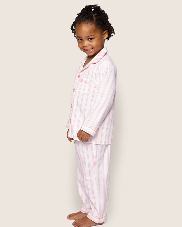 A young child with curly hair smiles while wearing a Petite Plume Kids Twill Pajama Set in Pink and White Stripe, standing against a plain background.
