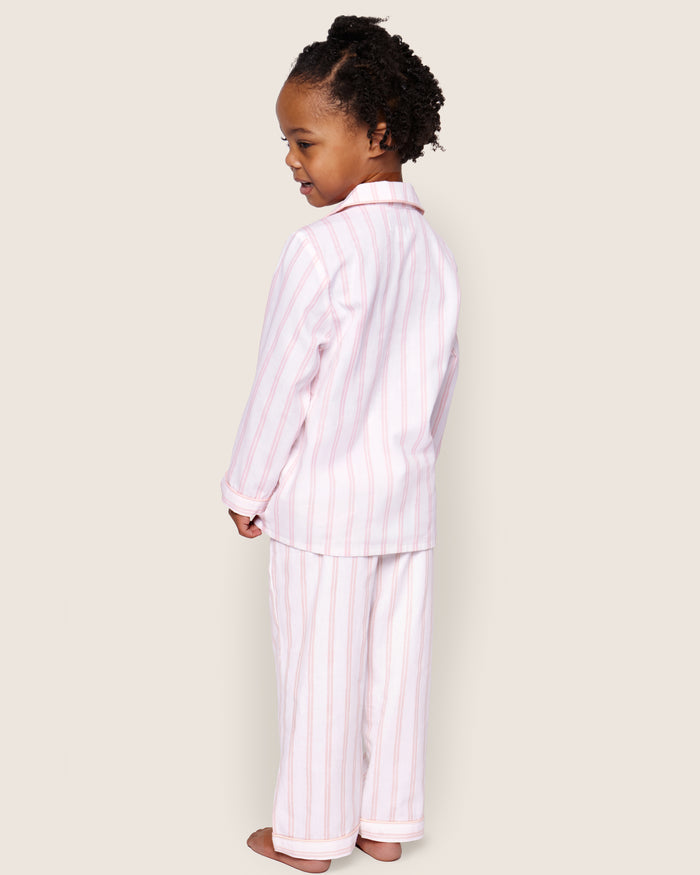 A curly-haired child smiles over their shoulder, wearing Petite Plumes Kids Twill Pajama Set in Pink and White Stripe. The cozy cotton sleepwear exudes charm against a light-colored background, enhancing the delightful scene.