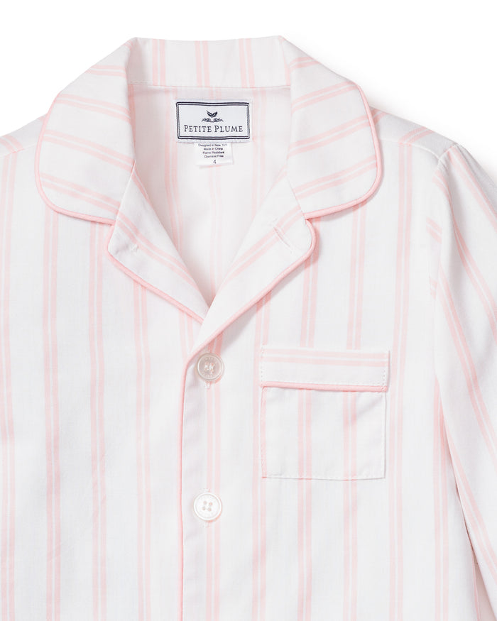 Close-up of a luxurious Kids Twill Pajama top in pink and white stripes. It has a notched collar, chest pocket, and two buttons. The label reads Petite Plume. The cotton fabric looks soft and smooth.