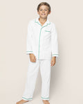 A smiling boy stands barefoot on a beige background, wearing Petite Plumes cozy Kids Twill Pajama Set in white with green piping. The button-up shirt includes a pocket and is paired with matching pants.