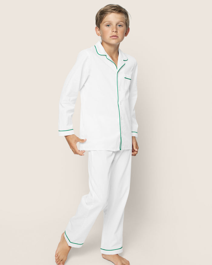 A boy stands barefoot on a plain background, wearing the Petite Plume Kids Twill Pajama Set in White with Green Piping. This cotton sleepwear is flame-retardant, ensuring comfort and peaceful nights.