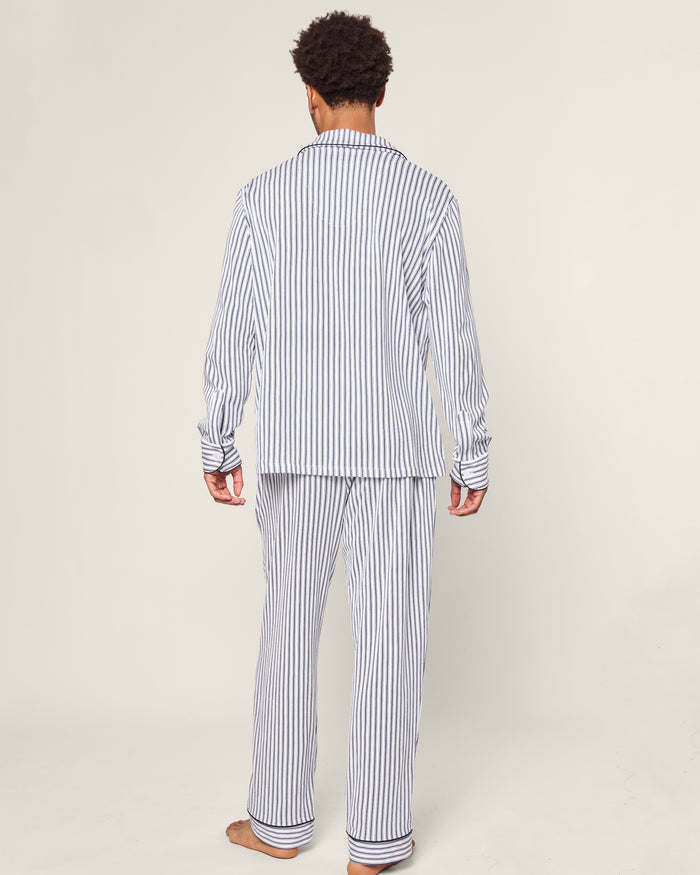 A person faces away from the camera, wearing the Mens Pima Pajama Set in Navy French Ticking by Petite Plume. The background is a plain, light color.