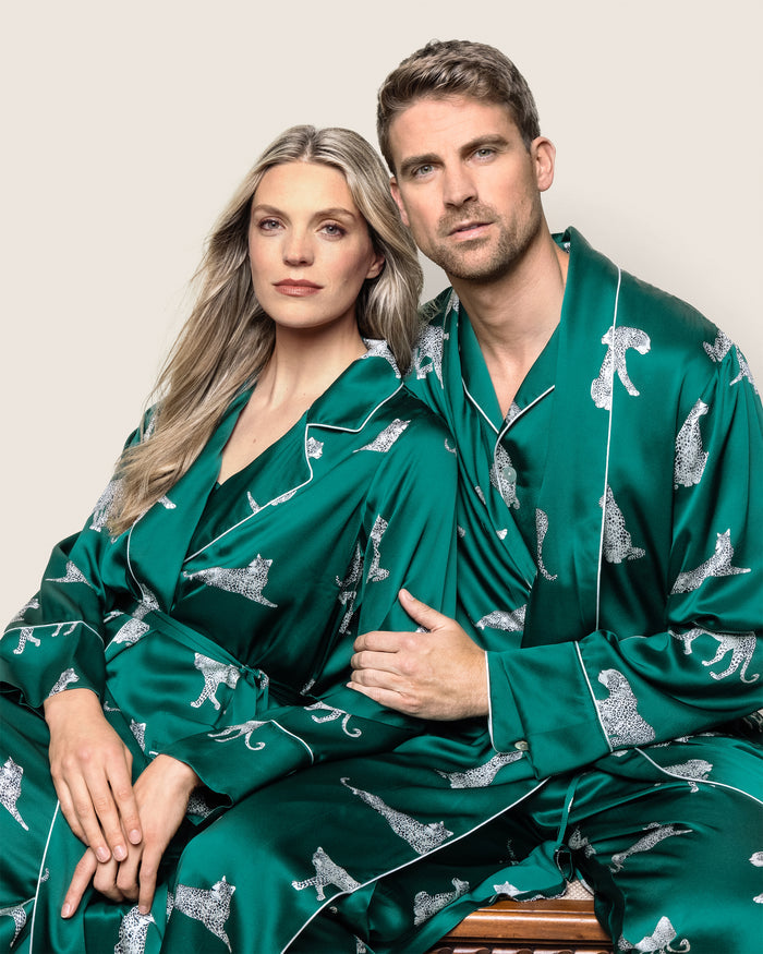 A woman and man sit closely, wrapped in matching green silk with a leopard print pattern. The man wears Petite Plumes Mens Silk Long Robe in Panthère de Luxe. Her hair is long and blonde, while his hair is short and light brown. They gaze directly at the camera against a plain background.