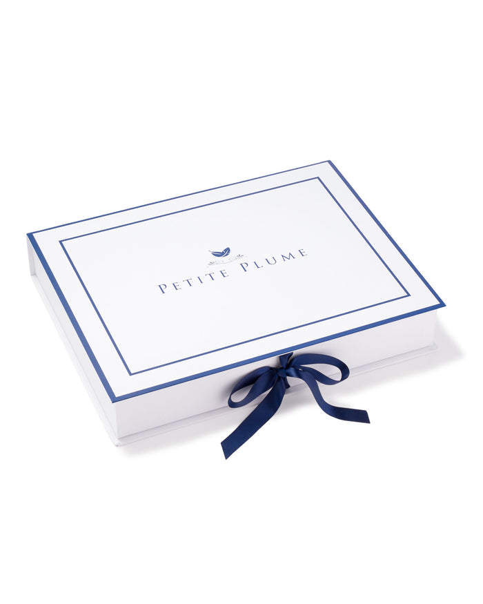 A white rectangular gift box features PETITE PLUME and a feather logo on the lid, outlined with blue lines and tied with a blue ribbon. It exudes elegance, hinting at luxurious items like the Mens Silk Pajama Set in Panthère de Luxe inside.