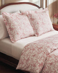 A neatly made bed features the Pink Timeless Toile Bedding Set with white sheets, a pink floral comforter, and two matching pillows against a curved, upholstered headboard in an elegant, classic bedroom.