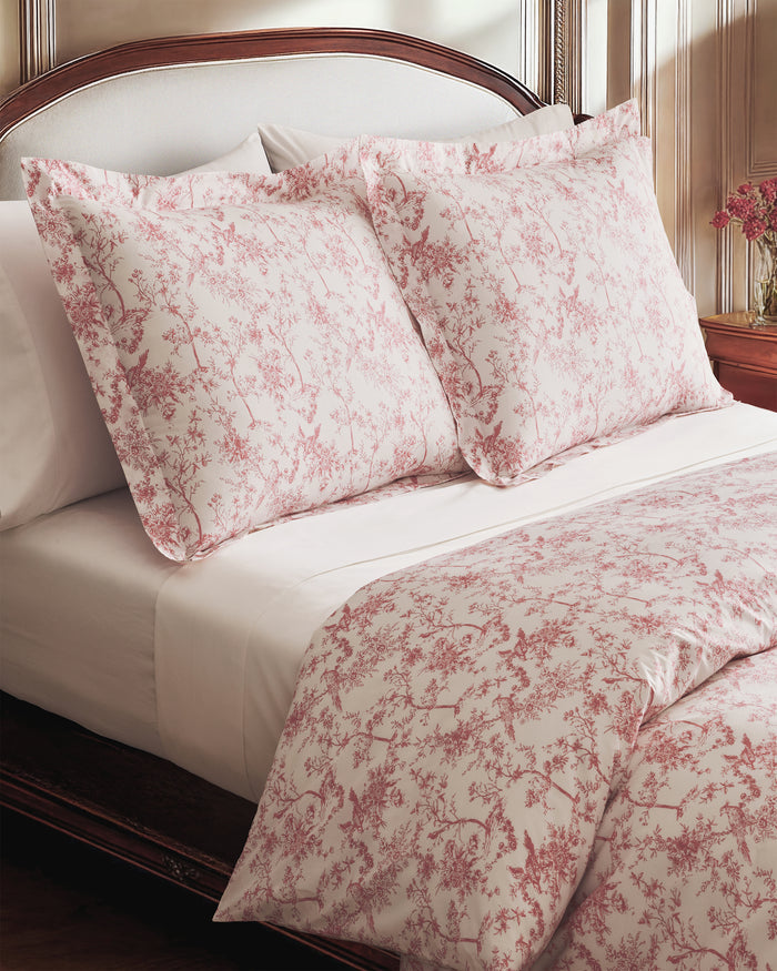 A neatly made bed features the Pink Timeless Toile Bedding Set with white sheets, a pink floral comforter, and two matching pillows against a curved, upholstered headboard in an elegant, classic bedroom.