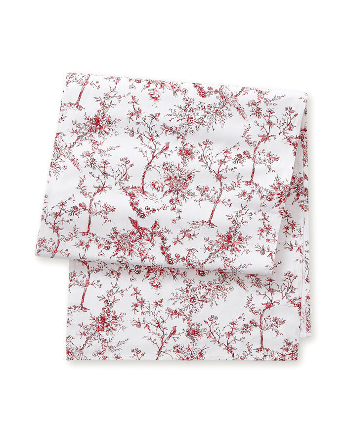 The Signature Twill Table Runner in Red Timeless Toile features a folded white fabric with a delicate red pattern of birds, trees, and branches.