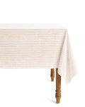The Signature Twill Tablecloth in Camel Ticking, featuring beige and white stripes, drapes over a wooden table with turned legs and is photographed against a plain white background.