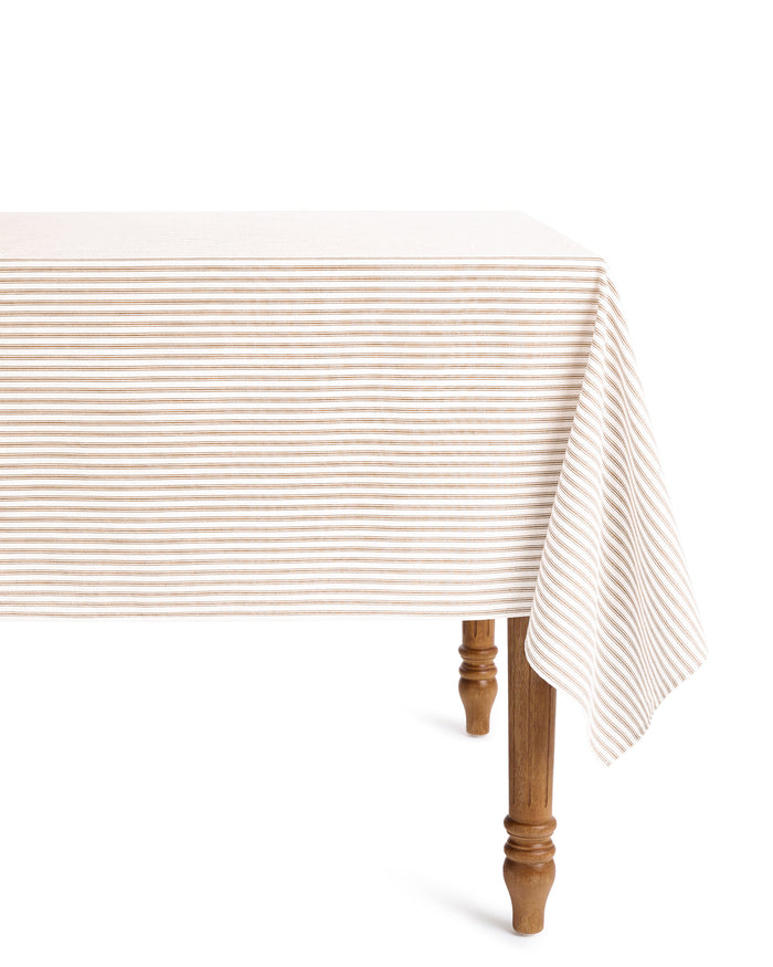 The Signature Twill Tablecloth in Camel Ticking, featuring beige and white stripes, drapes over a wooden table with turned legs and is photographed against a plain white background.