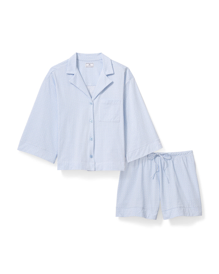 A woman with long curly hair models The Olivia Lightweight Pima Short Set in Periwinkle Geo—a light blue, long-sleeved button-up pajama top and matching dotted shorts—against a plain light background.