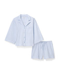 A woman with long curly hair models The Olivia Lightweight Pima Short Set in Periwinkle Geo—a light blue, long-sleeved button-up pajama top and matching dotted shorts—against a plain light background.
