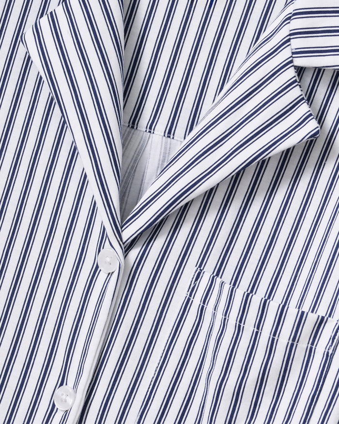 A person with long dark hair is wearing Petite Plumes Olivia Wide Leg Pima Pajama Set in Navy French Ticking, crafted from luxurious Peruvian Pima cotton. The button-up shirt and wide-legged pants have vertical stripes in alternating dark and light colors. They stand barefoot against a plain background.