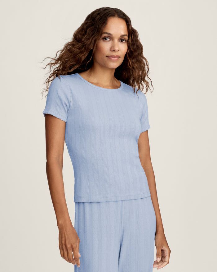 A woman with long wavy hair wears the Women's Pointelle T-Shirt in Periwinkle and matching pants, standing against a plain light background and looking at the camera with a slight smile.