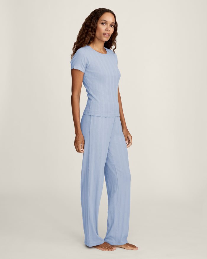 A woman with long wavy hair wears the Women's Pointelle T-Shirt in Periwinkle and matching pants, standing against a plain light background and looking at the camera with a slight smile.