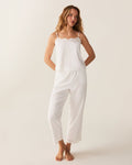 A woman with wavy hair stands barefoot on a light background, wearing the Women's Voile Fleur Cami Pant Set in White—featuring a sleeveless top and pants with lace trim. Her arms are behind her back.