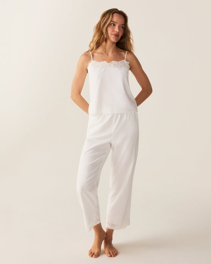 A woman with wavy hair stands barefoot on a light background, wearing the Women's Voile Fleur Cami Pant Set in White—featuring a sleeveless top and pants with lace trim. Her arms are behind her back.
