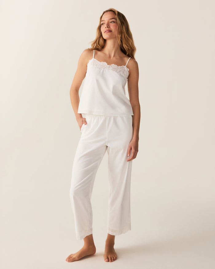 A woman with wavy hair stands barefoot on a light background, wearing the Women's Voile Fleur Cami Pant Set in White—featuring a sleeveless top and pants with lace trim. Her arms are behind her back.