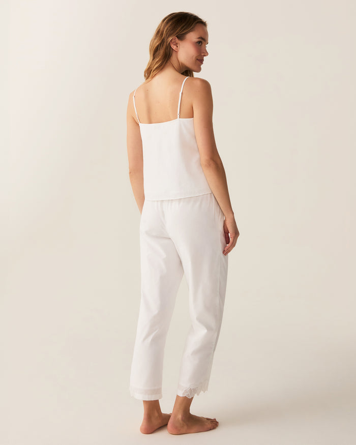 A woman with wavy hair stands barefoot on a light background, wearing the Women's Voile Fleur Cami Pant Set in White—featuring a sleeveless top and pants with lace trim. Her arms are behind her back.