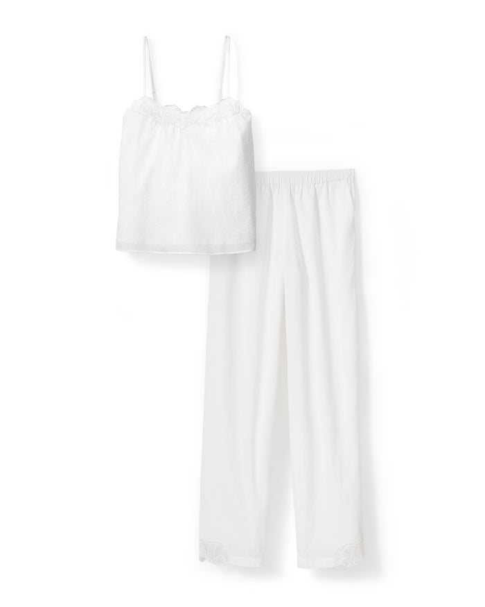 A woman with wavy hair stands barefoot on a light background, wearing the Women's Voile Fleur Cami Pant Set in White—featuring a sleeveless top and pants with lace trim. Her arms are behind her back.