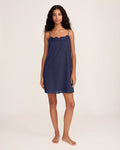 A woman with long curly hair wears the Women's Voile Fleur Slip in Navy, a sleeveless nightdress with lace trim at the neckline. She stands barefoot against a plain, light background, facing forward and smiling softly.
