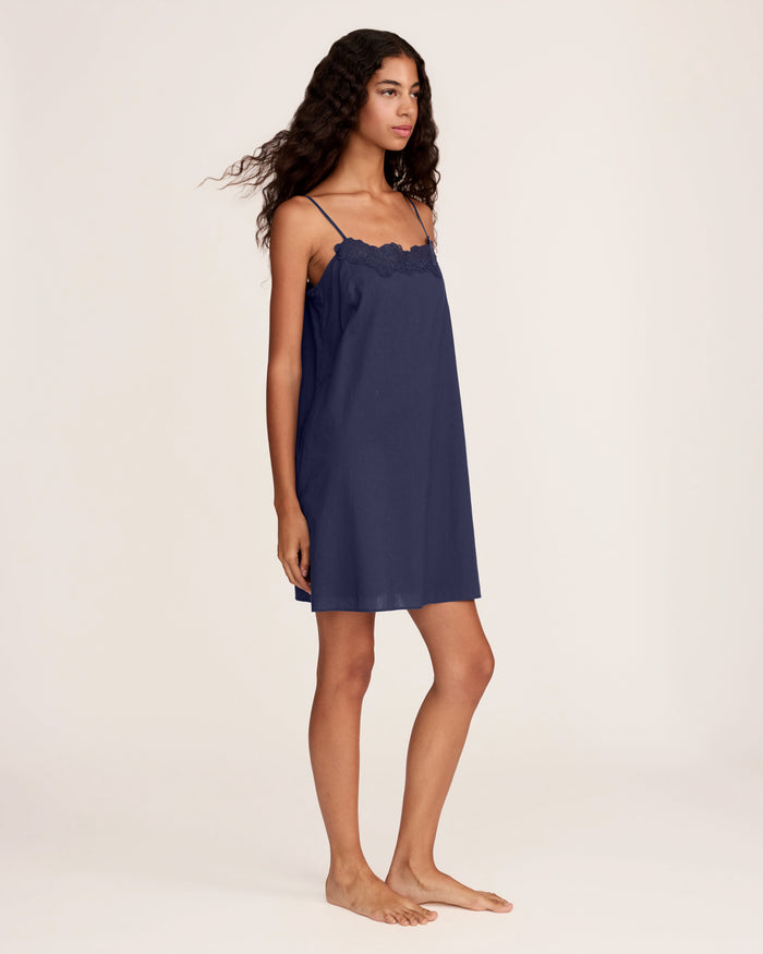 A woman with long curly hair wears the Women's Voile Fleur Slip in Navy, a sleeveless nightdress with lace trim at the neckline. She stands barefoot against a plain, light background, facing forward and smiling softly.