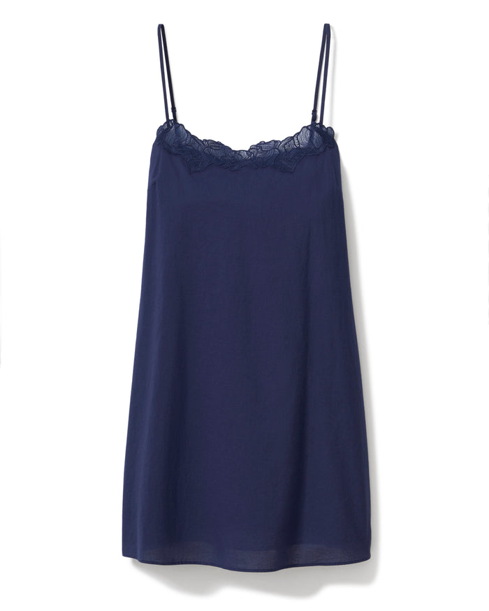 A woman with long curly hair wears the Women's Voile Fleur Slip in Navy, a sleeveless nightdress with lace trim at the neckline. She stands barefoot against a plain, light background, facing forward and smiling softly.