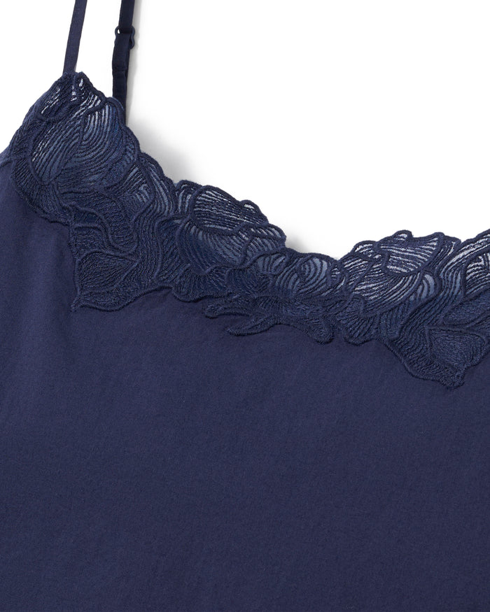 A woman with long curly hair wears the Women's Voile Fleur Slip in Navy, a sleeveless nightdress with lace trim at the neckline. She stands barefoot against a plain, light background, facing forward and smiling softly.