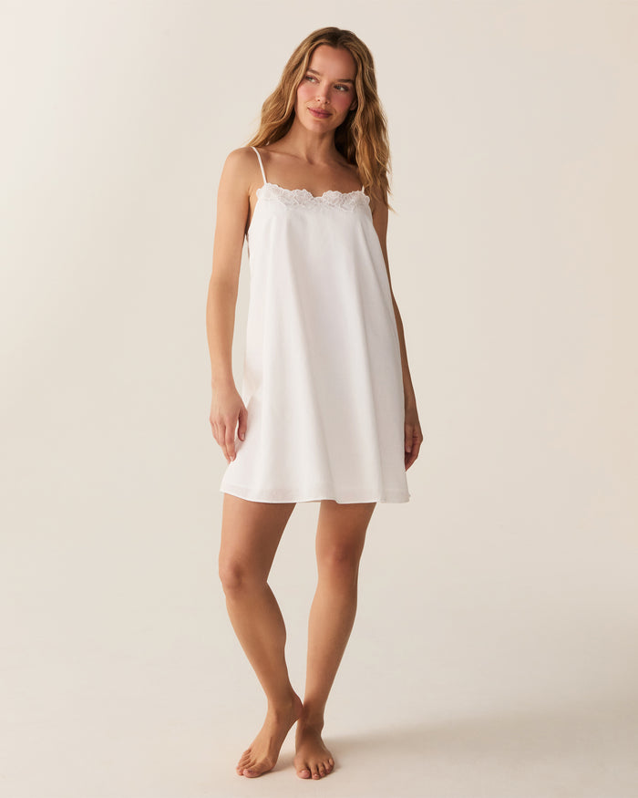 A woman with long, wavy hair stands barefoot against a plain light background, looking at the camera with a slight smile while wearing the Women's Voile Fleur Slip in White.