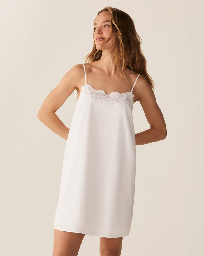 A woman with long, wavy hair stands barefoot against a plain light background, looking at the camera with a slight smile while wearing the Women's Voile Fleur Slip in White.